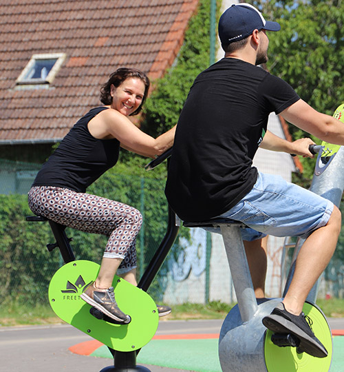 une femme sourit à un homme tous les deux sur des vélos de fitness en extérieur.