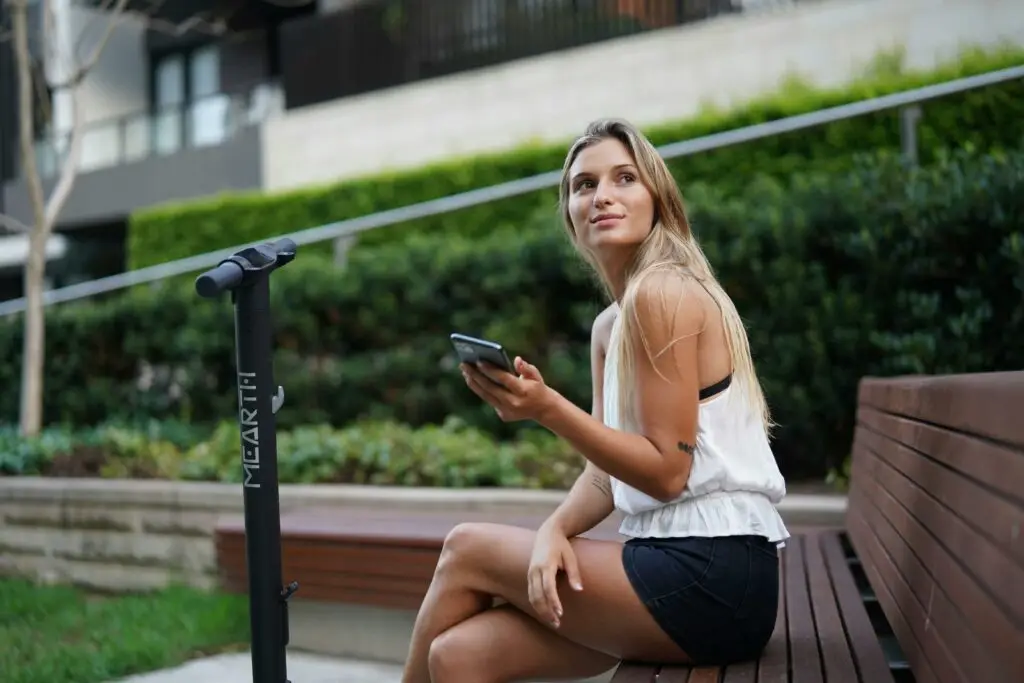 femme souriante avec un portable dans la main et une trottinette devant elle.